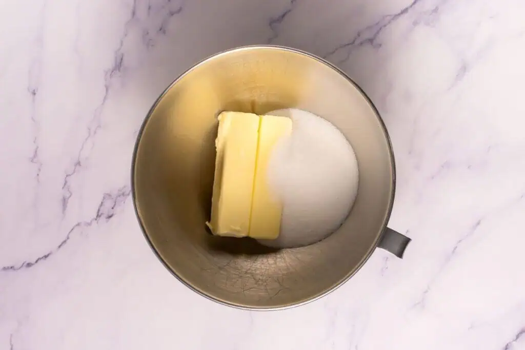 A metal mixing bowl containing two sticks of butter and a mound of granulated sugar on a marble surface.