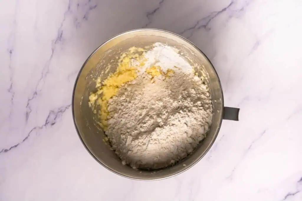 A mixing bowl containing creamed butter and sugar with flour added on top, set on a white marble surface.