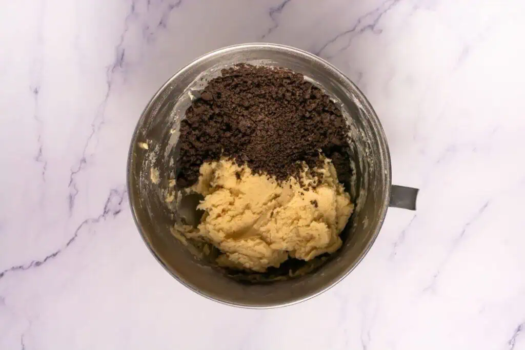 A metal mixing bowl containing pale cookie dough and crushed chocolate cookie pieces on a white marble surface.