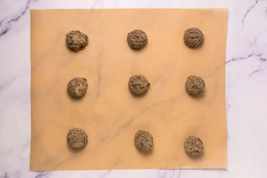Nine scoops of cookie dough are evenly spaced on a sheet of parchment paper placed on a marble surface.