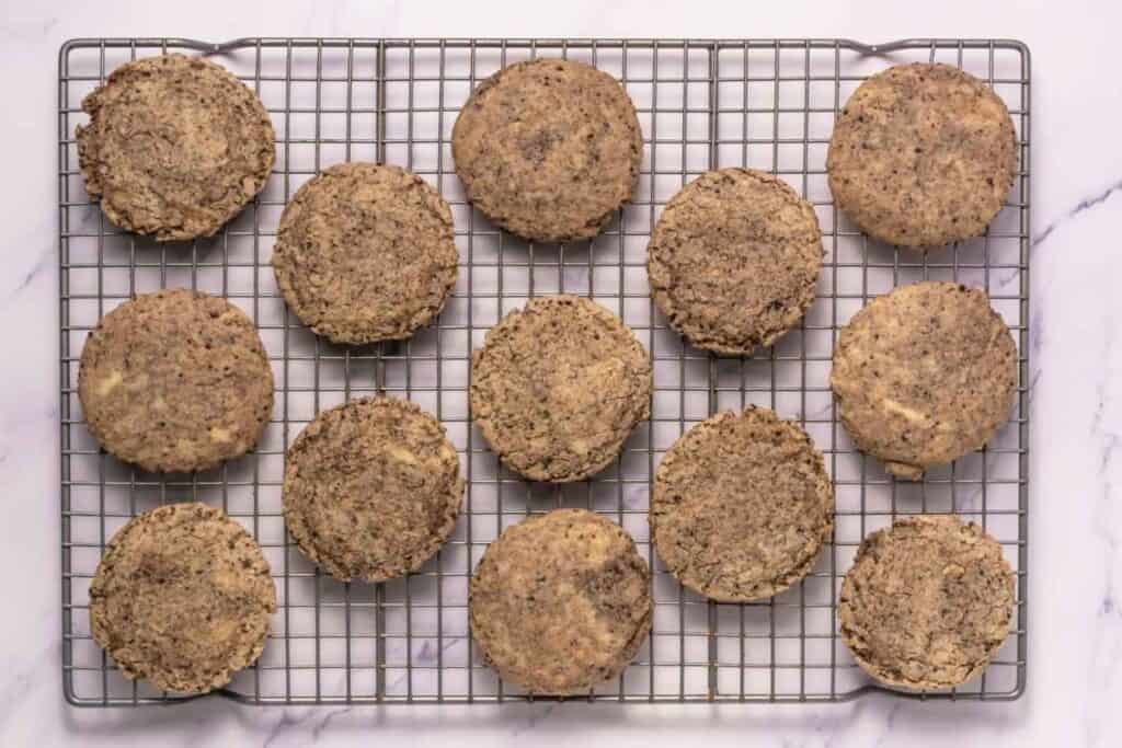 Fourteen round cookies with a speckled appearance are arranged on a wire cooling rack placed on a white surface.