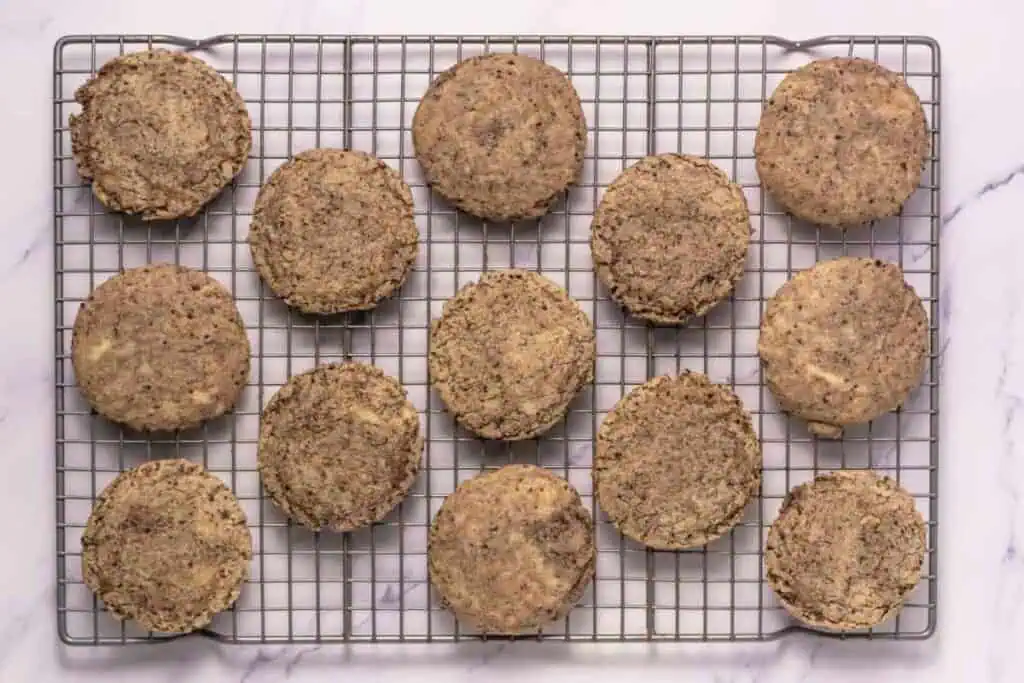 Fourteen round cookies with a speckled appearance are arranged on a wire cooling rack placed on a white surface.