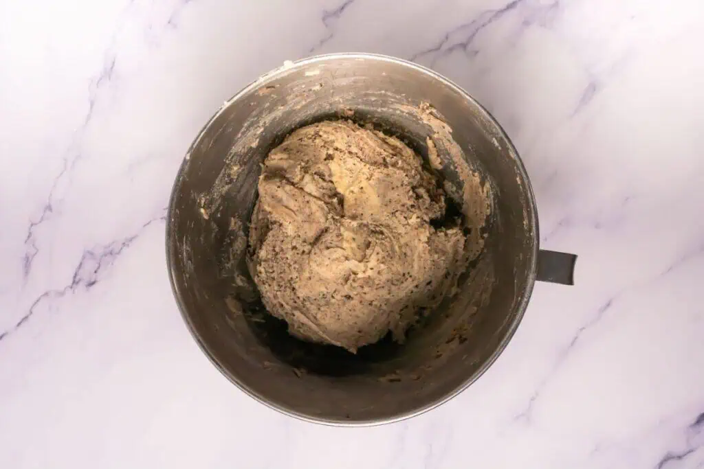 A metal mixing bowl containing a large amount of raw cookie dough sits on a white marble countertop.