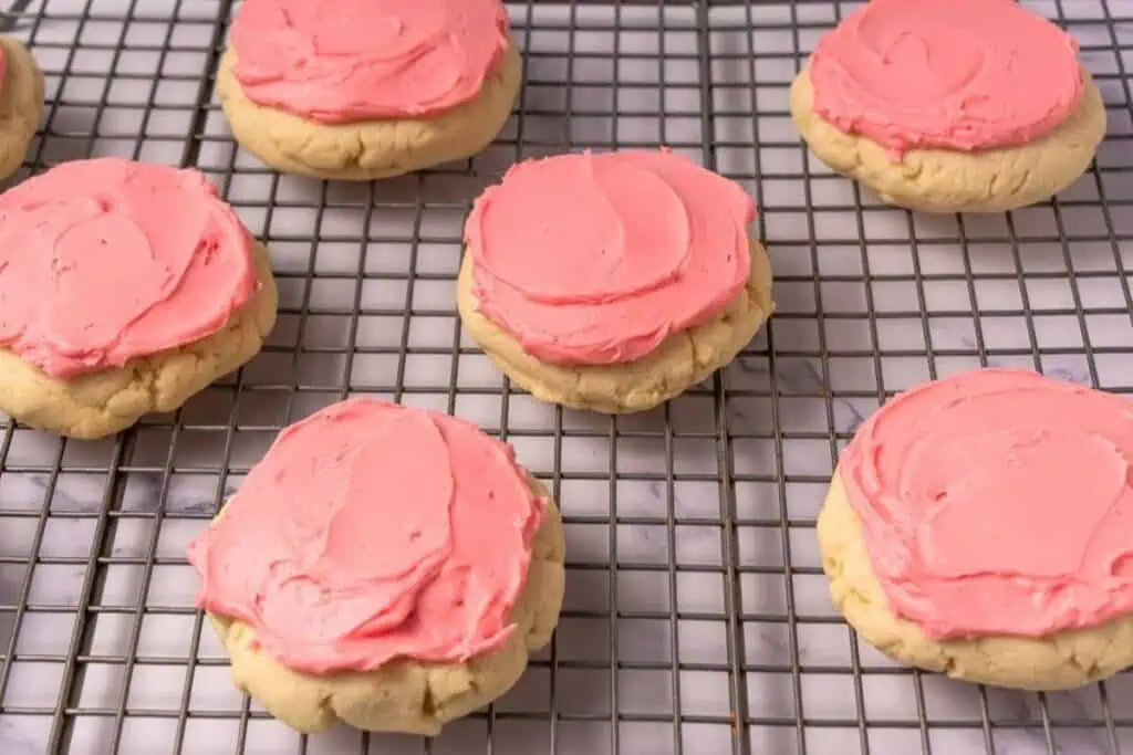Sugar cookies with pink frosting are cooling on a metal wire rack placed on a marble surface.
