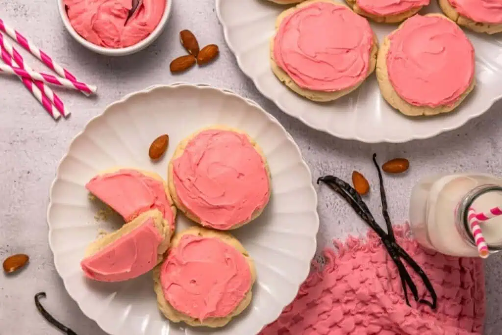 A plate of sugar cookies with pink frosting, a small bowl of extra frosting, almonds, vanilla beans, a glass of milk with a striped straw, and pink napkin on a white surface.