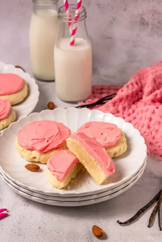 Three sugar cookies with pink frosting on a white plate, one cookie cut in half, with glasses of milk and a pink cloth in the background.