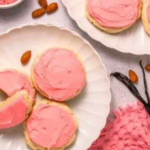 Two plates of round sugar cookies with pink frosting, some with bites taken out, are surrounded by almonds, vanilla beans, a bottle of milk with a straw, and pink napkins.