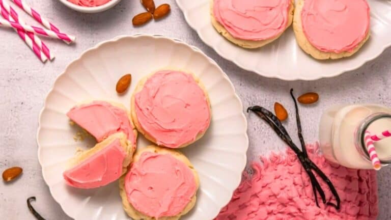 Two plates of round sugar cookies with pink frosting, some with bites taken out, are surrounded by almonds, vanilla beans, a bottle of milk with a straw, and pink napkins.