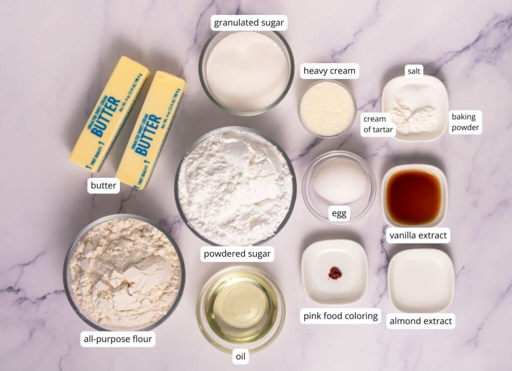 Ingredients for baking arranged on a marble surface, including butter, flour, powdered sugar, oil, granulated sugar, heavy cream, egg, extracts, food coloring, salt, cream of tartar, and baking powder.