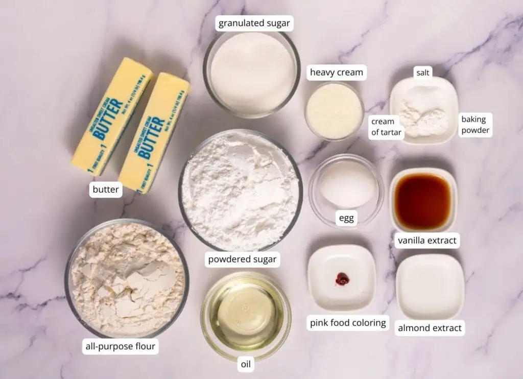 Ingredients for baking arranged on a marble surface, including butter, flour, powdered sugar, oil, granulated sugar, heavy cream, egg, extracts, food coloring, salt, cream of tartar, and baking powder.