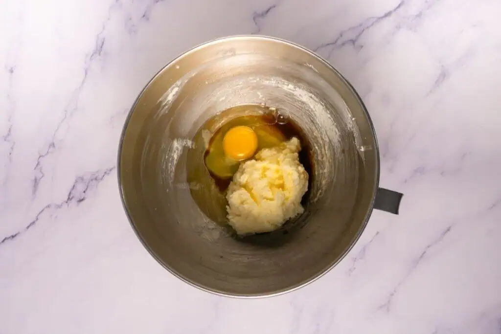 A metal mixing bowl contains creamed butter and sugar, an egg, and vanilla extract on a marble countertop.