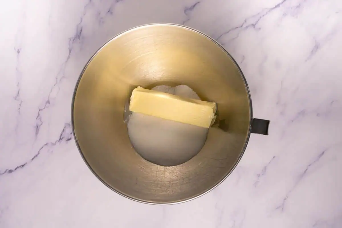 A metal mixing bowl containing a stick of butter and granulated sugar on a light marble surface.