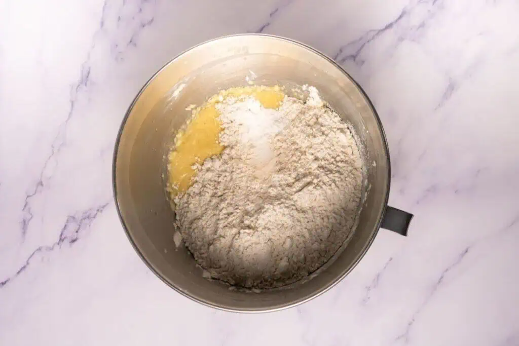 A metal mixing bowl containing flour and a yellow batter mixture sits on a white marble surface.