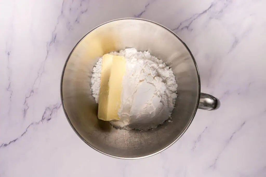 A metal mixing bowl containing a stick of butter and a mound of powdered sugar on a marble countertop.