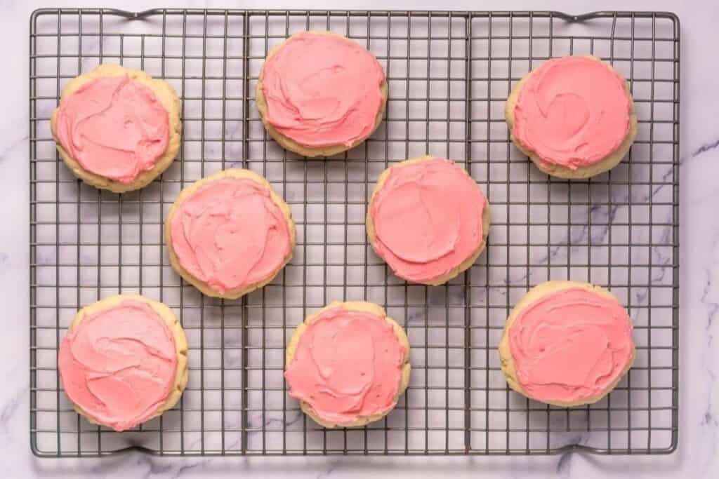 Eight sugar cookies with pink frosting are arranged on a wire cooling rack over a marble surface.