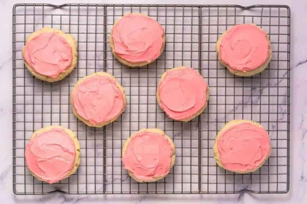 Eight sugar cookies with pink frosting are arranged on a wire cooling rack over a marble surface.