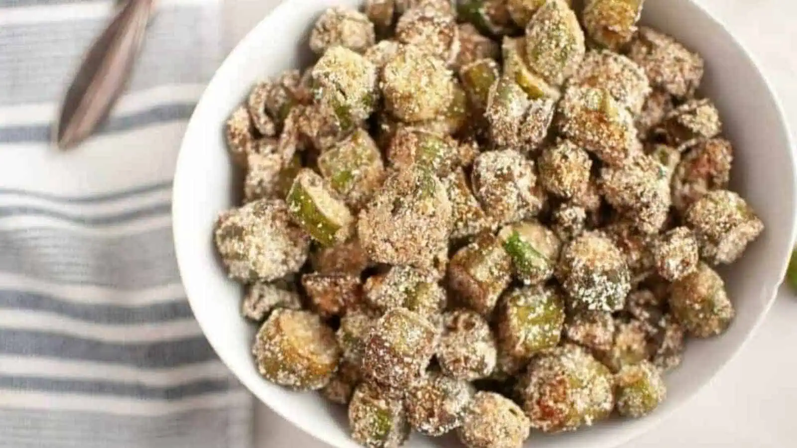 A white bowl filled with crispy, breaded and fried okra pieces, placed on a light surface next to a striped cloth napkin.
