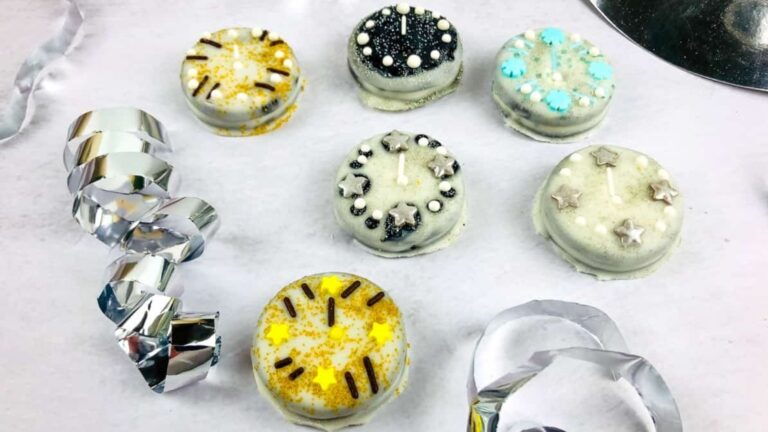Six decorated round cookies with white icing and various gold, black, and blue sprinkles are arranged on a white surface with silver ribbon decorations nearby.