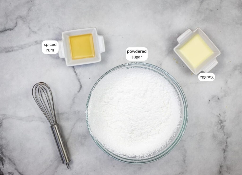 A bowl of powdered sugar, a dish of spiced rum, a dish of eggnog, and a whisk are arranged on a marble surface. Labels identify each ingredient.