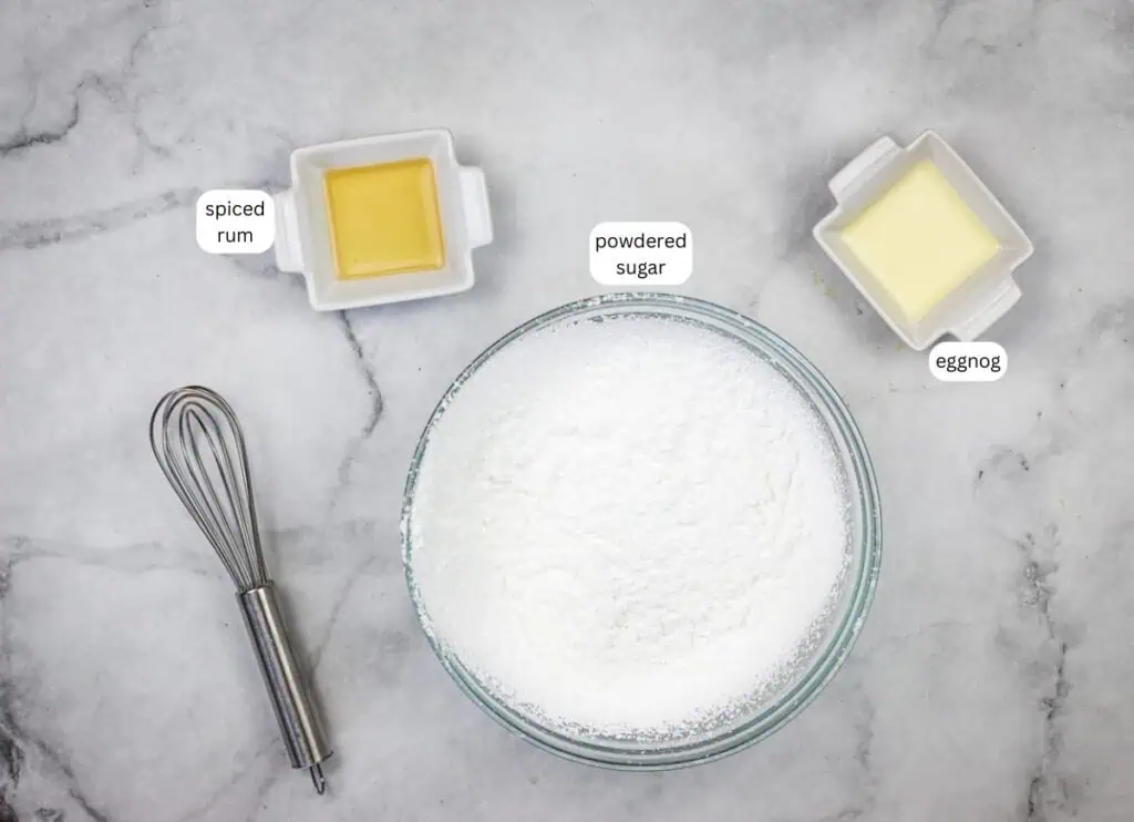 A bowl of powdered sugar, a dish of spiced rum, a dish of eggnog, and a whisk are arranged on a marble surface. Labels identify each ingredient.