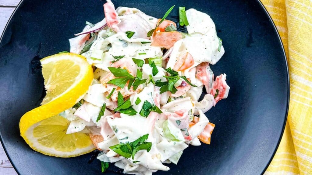 A creamy salad with crab sticks, chopped vegetables, and fresh herbs, garnished with two lemon slices, served on a black plate.