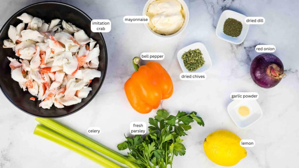 Ingredients for a deli seafood salad are laid out on a marble surface, including imitation crab, mayonnaise, bell pepper, dried dill, red onion, celery, dried chives, garlic powder, fresh parsley, and lemon.