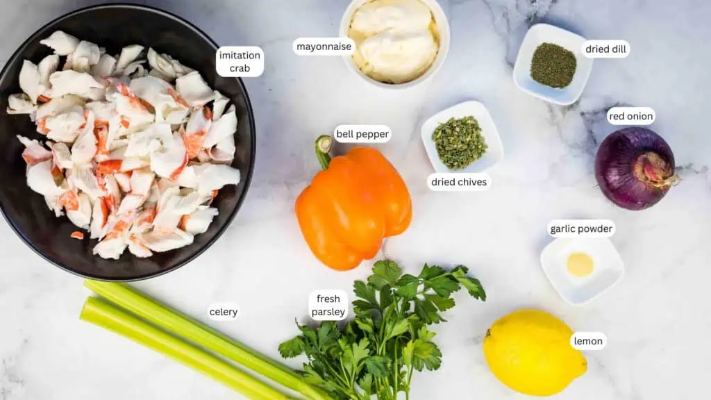 Ingredients for a deli seafood salad are laid out on a marble surface, including imitation crab, mayonnaise, bell pepper, dried dill, red onion, celery, dried chives, garlic powder, fresh parsley, and lemon.