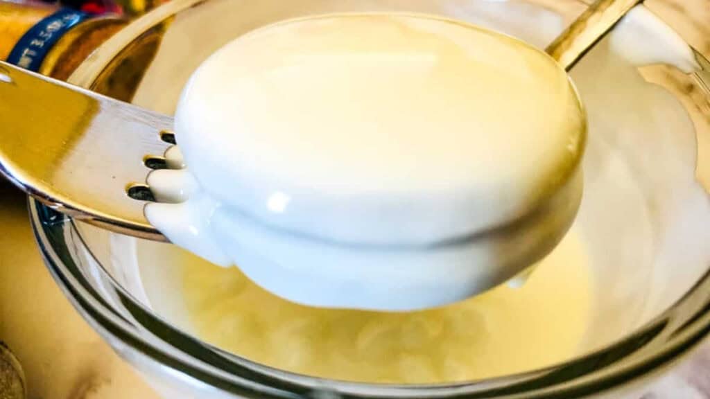 A metal ladle holds a thick, creamy white liquid over a glass bowl, with some of the liquid dripping back into the bowl.