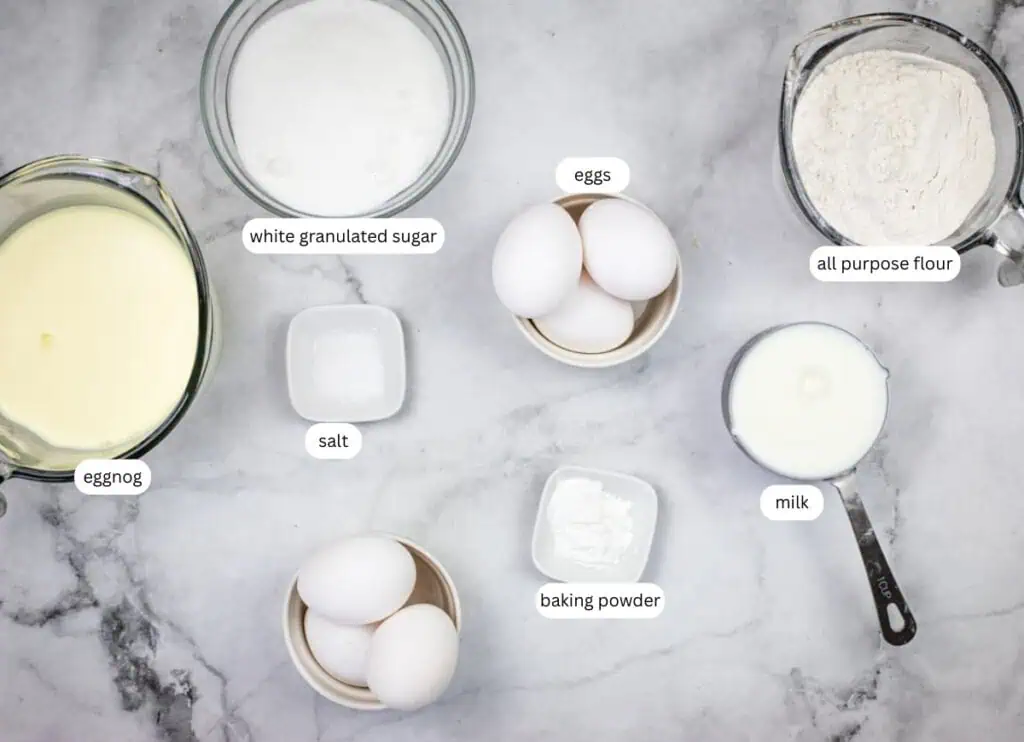 Labeled ingredients for baking on a marble surface: eggnog, sugar, eggs, salt, baking powder, all-purpose flour, and milk.