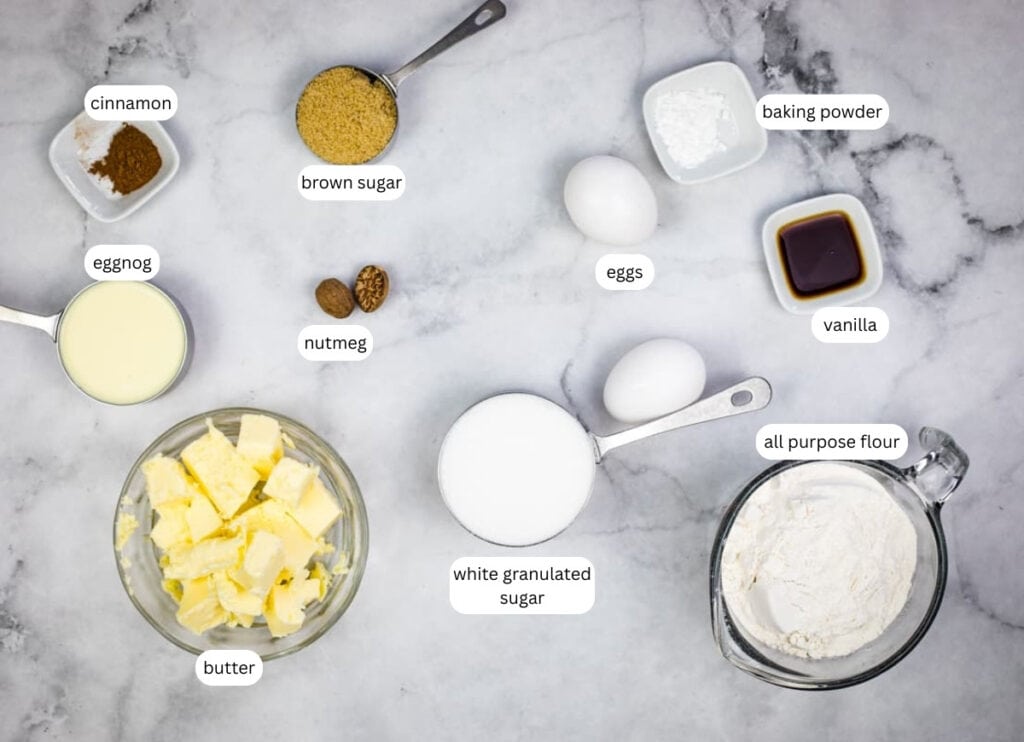 Various baking ingredients labeled on a marble surface, including butter, eggnog, brown sugar, nutmeg, cinnamon, eggs, white sugar, all-purpose flour, baking powder, and vanilla.