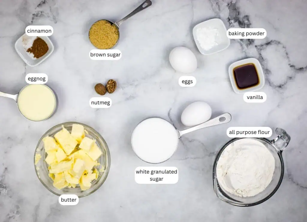 Various baking ingredients labeled on a marble surface, including butter, eggnog, brown sugar, nutmeg, cinnamon, eggs, white sugar, all-purpose flour, baking powder, and vanilla.