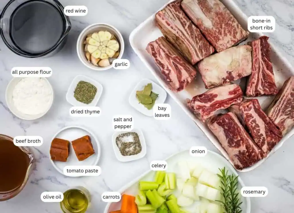 Ingredients for braised short ribs arranged on a surface, including bone-in short ribs, vegetables, herbs, beef broth, tomato paste, olive oil, flour, and red wine, each labeled.