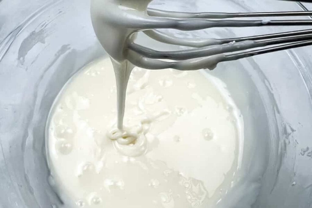 A metal whisk is lifting thick, smooth white batter from a glass bowl.