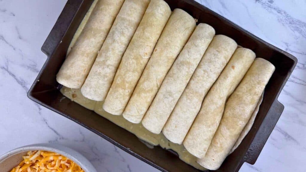 A baking dish filled with rolled tortillas placed side by side, next to a bowl of shredded cheese on a marble countertop.