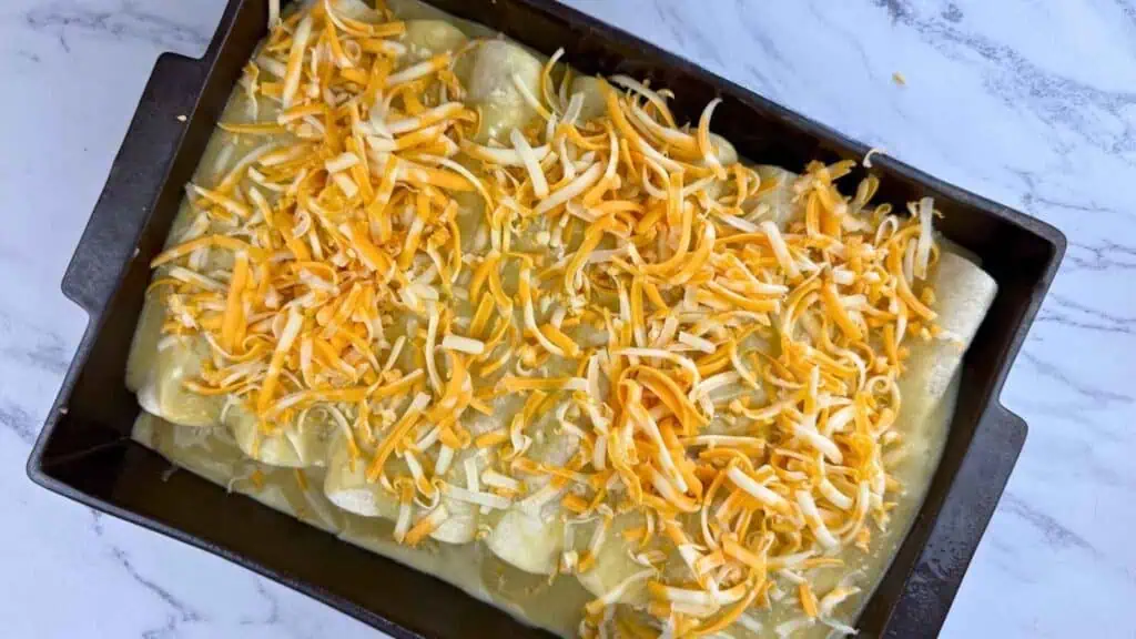 A rectangular baking dish filled with rolled tortillas covered in shredded yellow and white cheese on a marble countertop.