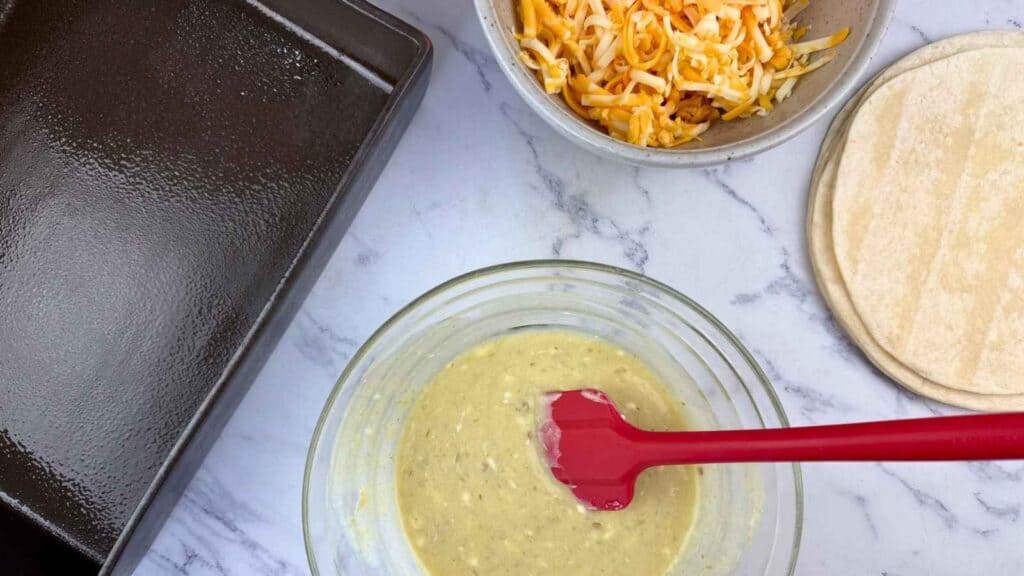 A glass bowl with batter and a red spatula, a bowl of shredded cheese, corn tortillas, and a greased baking pan on a marble countertop.