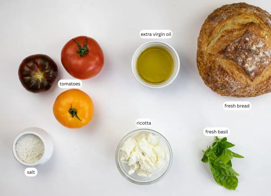 Three tomatoes, a bowl of ricotta, a bowl of olive oil, a loaf of bread, a bowl of salt, and fresh basil leaves arranged on a white surface.