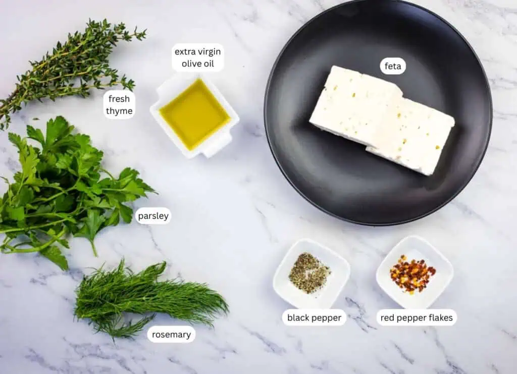 A black plate with feta cheese, and small dishes of black pepper, red pepper flakes, and olive oil, surrounded by sprigs of parsley, rosemary, and fresh thyme on a marble surface.