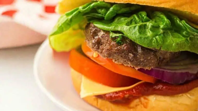 Close-up of a hamburger with lettuce, tomato, onion, cheese, ketchup, and a beef patty on a bun, served on a white plate.