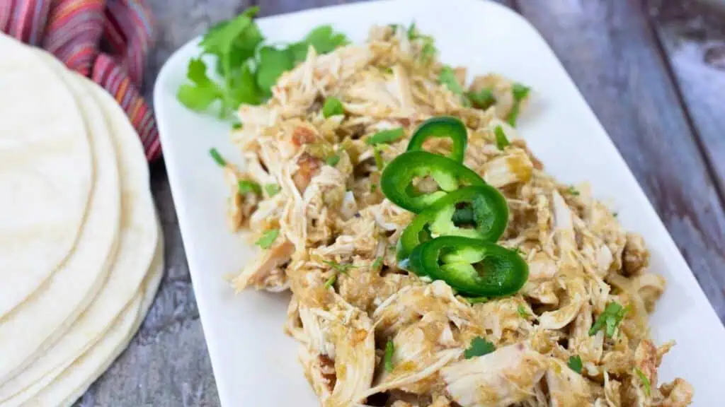 Shredded chicken with green sauce and sliced jalapeños on a white plate, garnished with cilantro, with tortillas nearby.