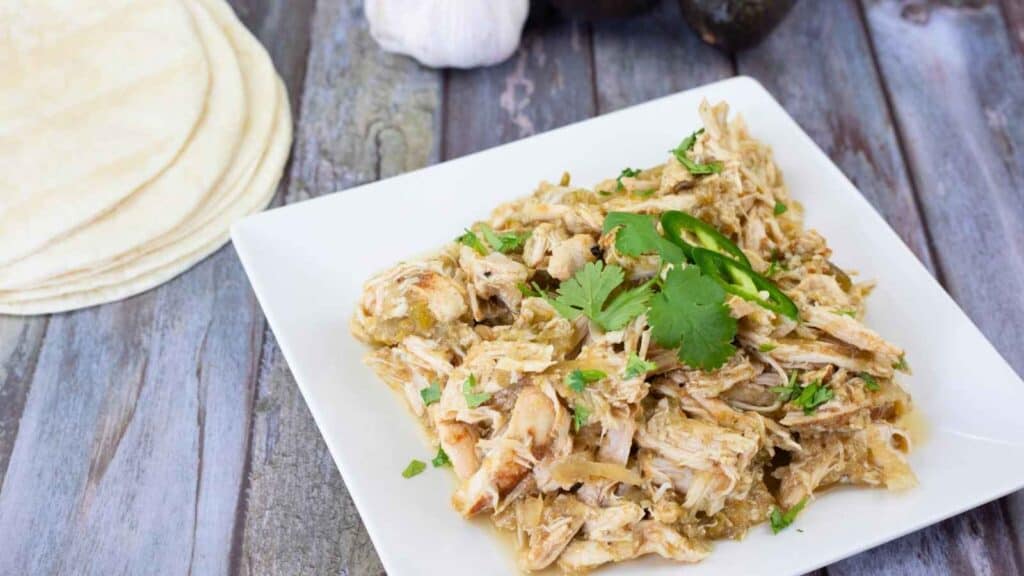 A white plate of shredded chicken garnished with sliced jalape&ntilde;o and cilantro sits next to a stack of tortillas on a wooden surface.