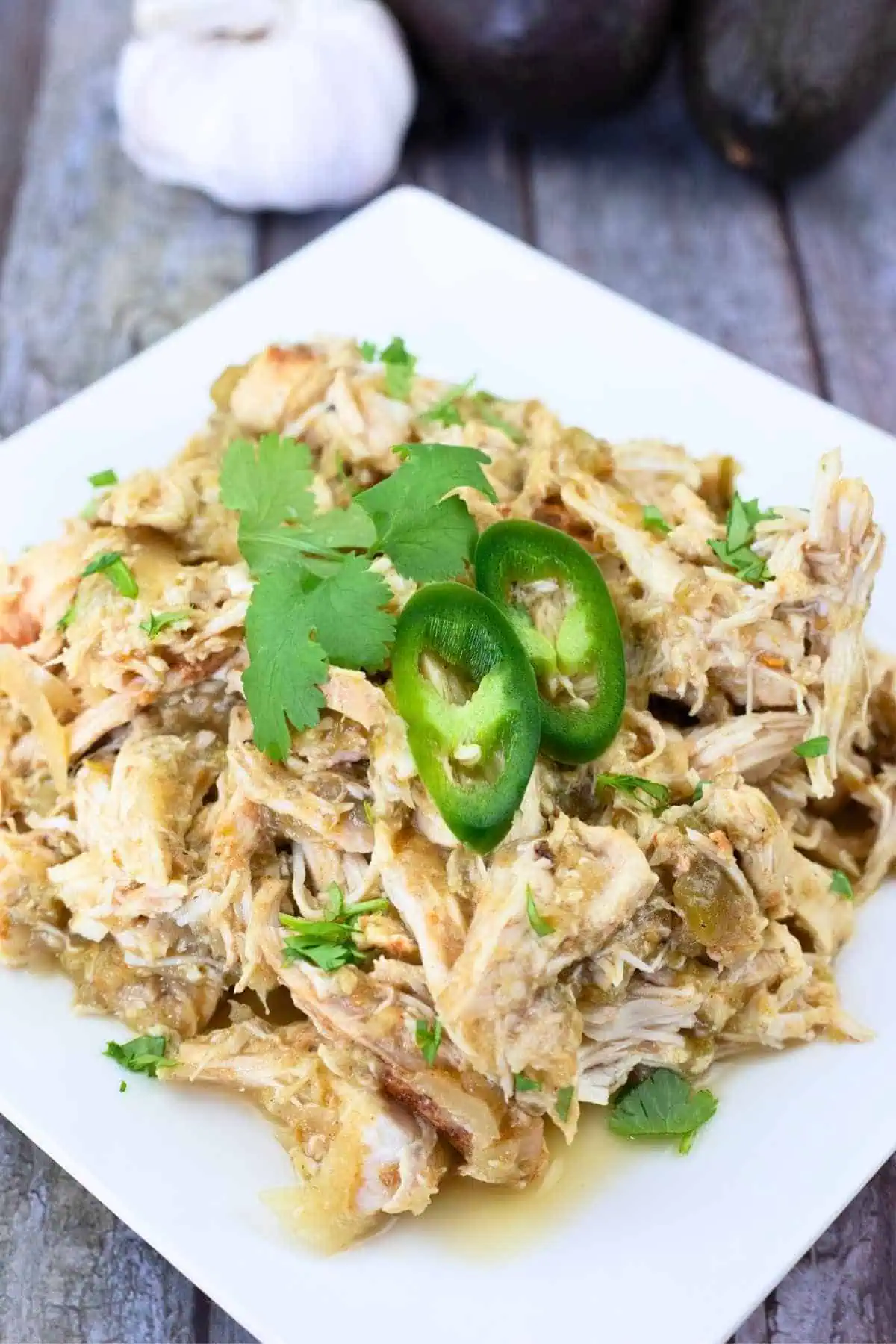 A plate of shredded chicken topped with sliced jalapeños and cilantro, served on a white square dish with garlic and avocados in the background.