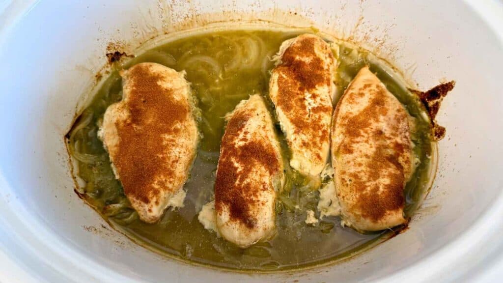 Four seasoned chicken breasts in a slow cooker with green sauce and sliced onions.