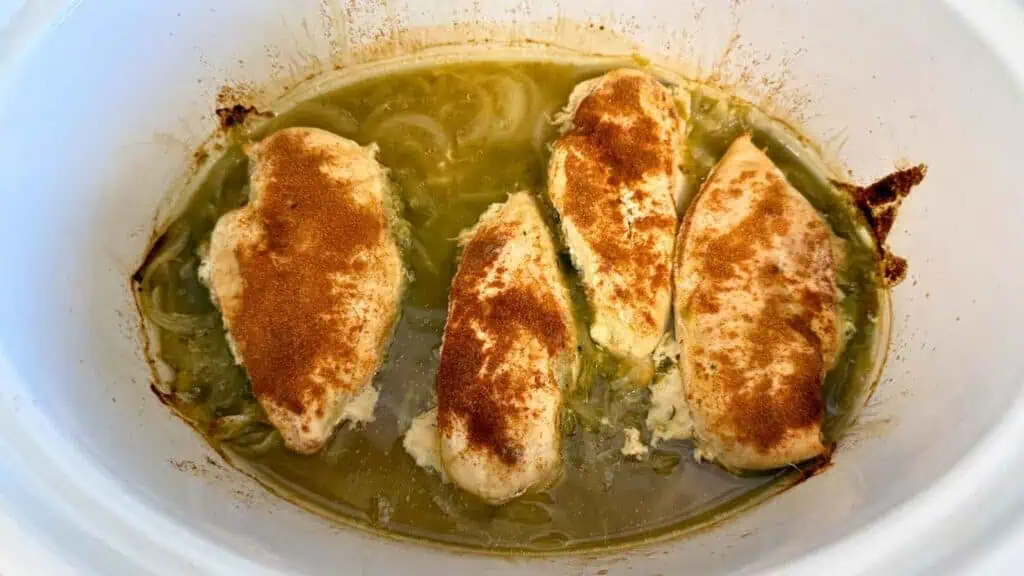 Four seasoned chicken breasts in a slow cooker with green sauce and sliced onions.