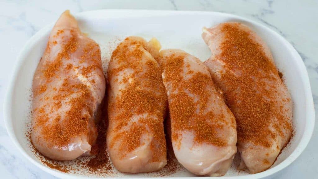 Four raw chicken breasts in a white dish, sprinkled with a reddish-brown seasoning, on a marble countertop.