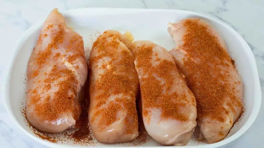 Four raw chicken breasts in a white dish, sprinkled with a reddish-brown seasoning, on a marble countertop.