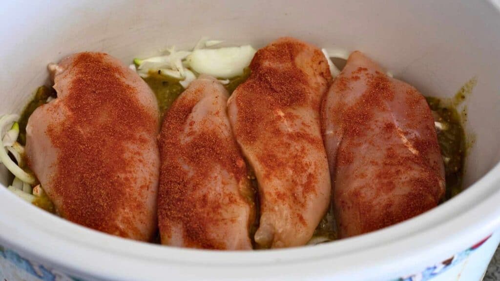 Four raw chicken breasts coated with seasoning are arranged in a slow cooker on top of sliced onions and green sauce.