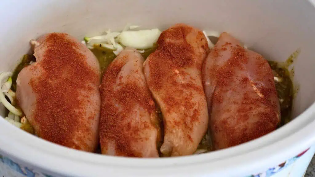 Four raw chicken breasts coated with seasoning are arranged in a slow cooker on top of sliced onions and green sauce.