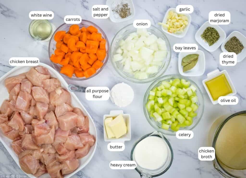 Ingredients for a chicken stew recipe arranged on a marble surface, including diced chicken, vegetables, herbs, butter, flour, olive oil, cream, wine, and chicken broth.
