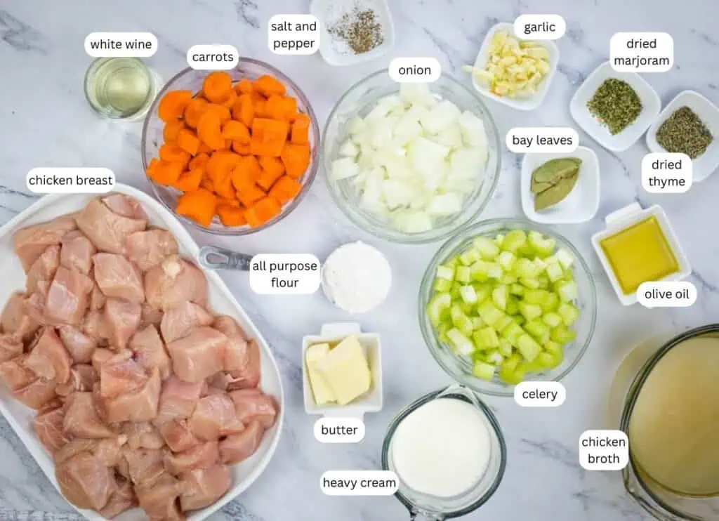 Ingredients for a chicken stew recipe arranged on a marble surface, including diced chicken, vegetables, herbs, butter, flour, olive oil, cream, wine, and chicken broth.